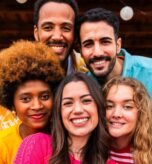 Multiethnic group of happy young friends having dinner barbecue party on rooftop at home - Multiracial cheerful young adult people having fun and bonding on a terrace balcony with city view, eating and drinking. Close-up portrait of funny faces.